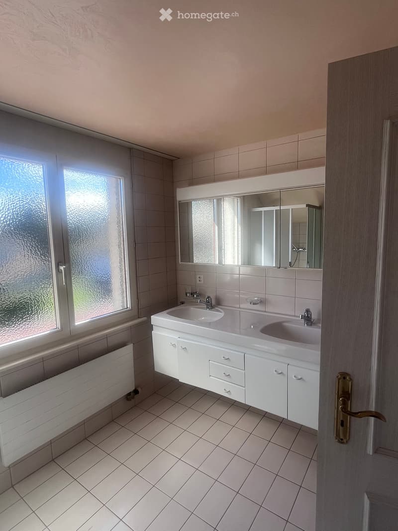 Modern bathroom with white tiled walls and floor, double sink vanity with mirrored cabinet, frosted glass window, and radiator below the window.