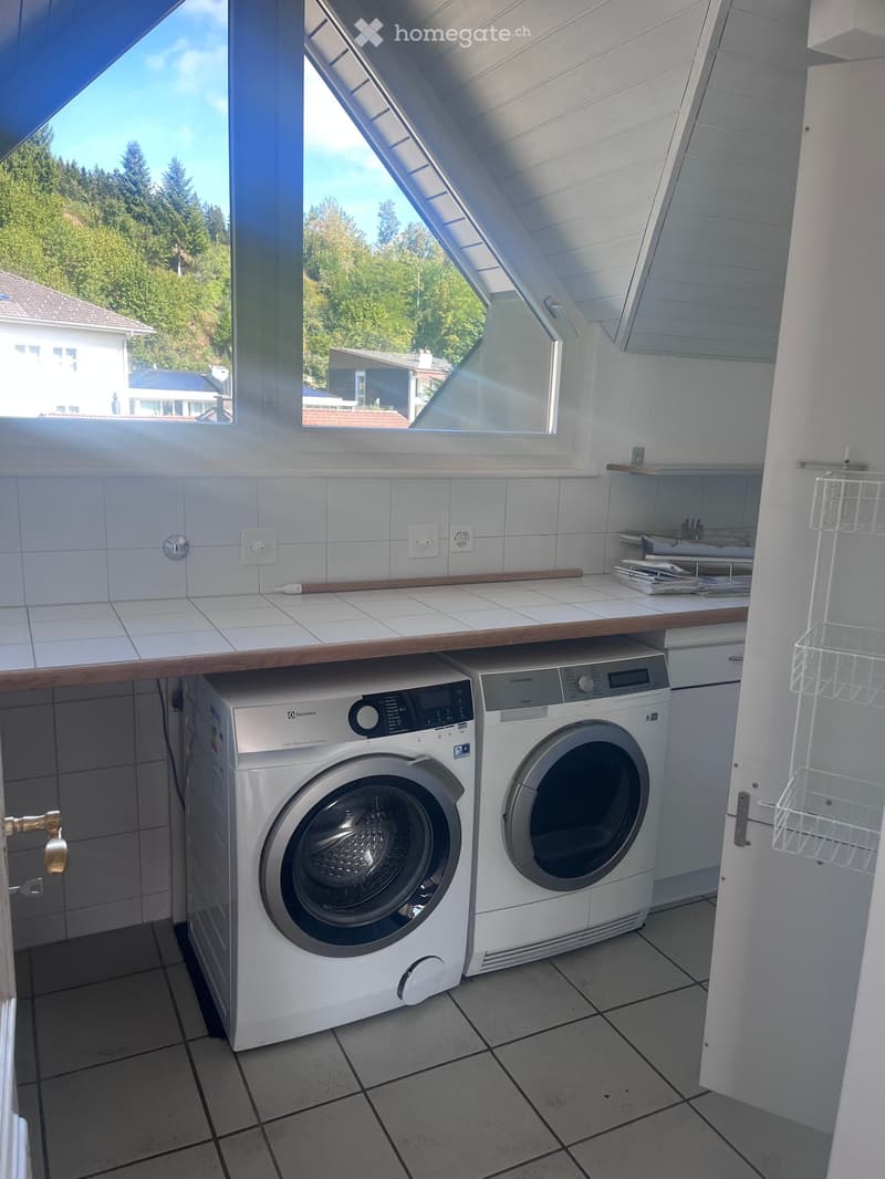 Bright, tiled laundry room with a washing machine and dryer under a wooden counter, with shelves and a window overlooking a green area.