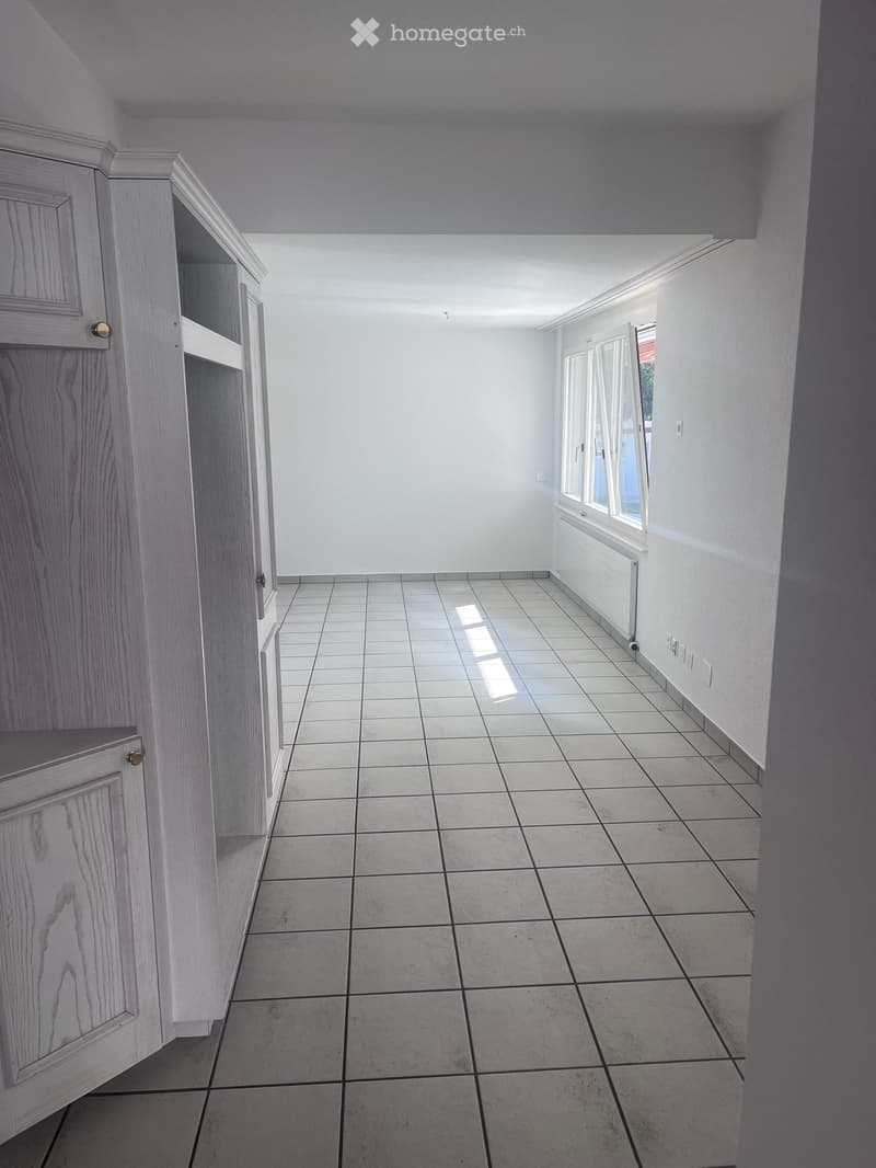 Empty hallway with tiled floor, white walls, and built-in wooden cabinetry on one side. A window at the far end allows natural light to enter.