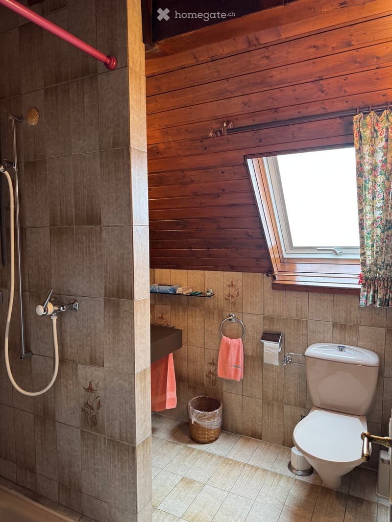 Bathroom with wooden walls, tiled shower area, toilet, sink, and window with floral curtain.