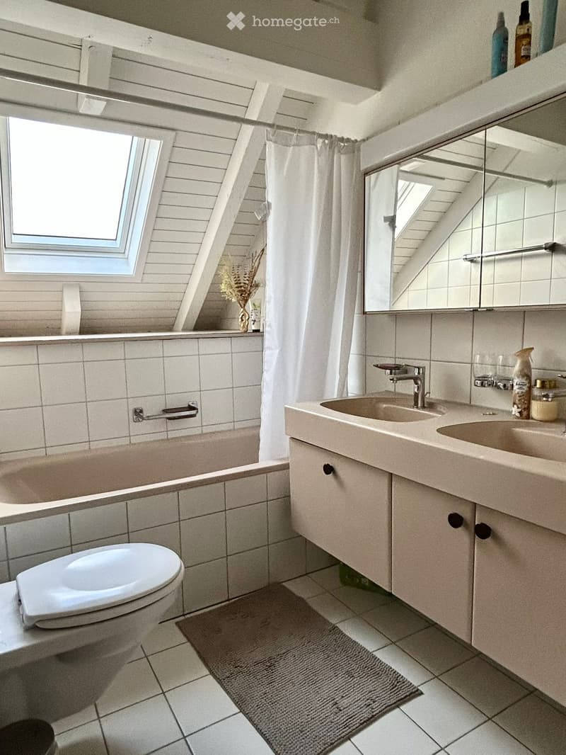 Modern bathroom with a bathtub, toilet, double sink, mirror, and a window with a shower curtain. Includes a small vase on the windowsill and a rug on the floor.