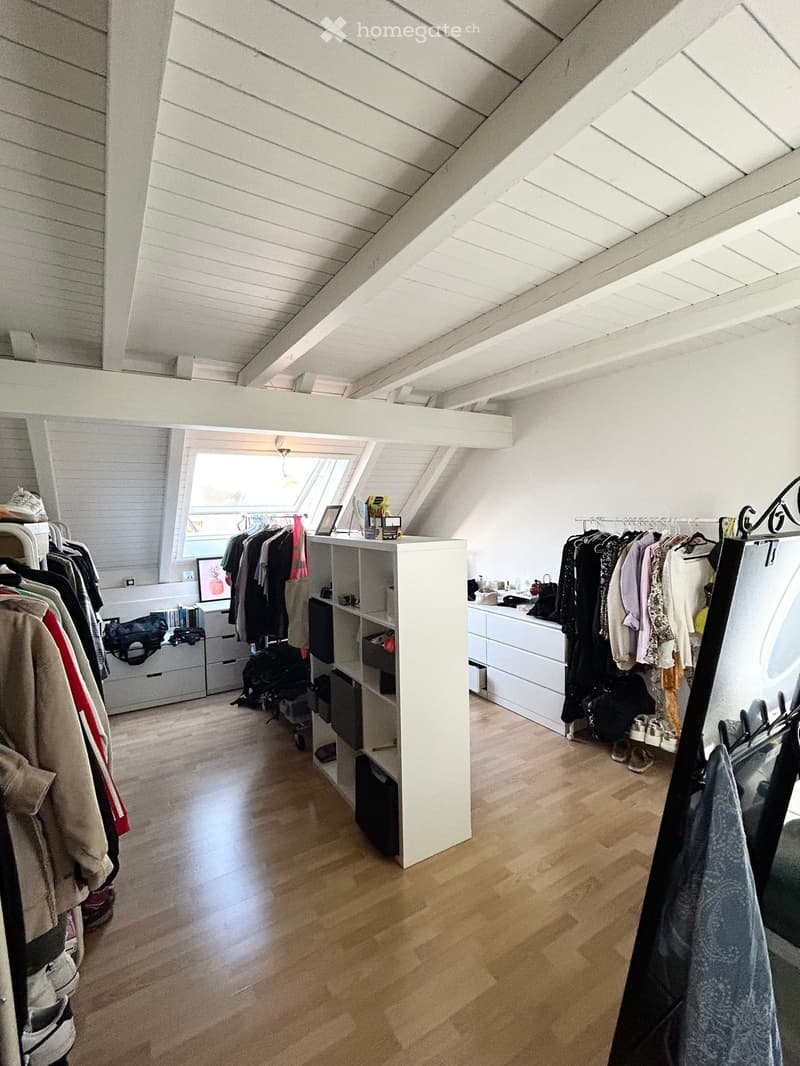 A well-lit attic room with wooden flooring, white walls, and exposed wooden beams. Features include a clothing rack, a shelf with various items, and a small table with a mirror.