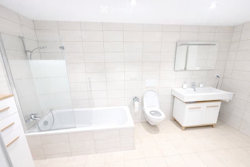 Modern bathroom with white tiles, a bathtub, toilet, sink, and a large mirror above the sink.