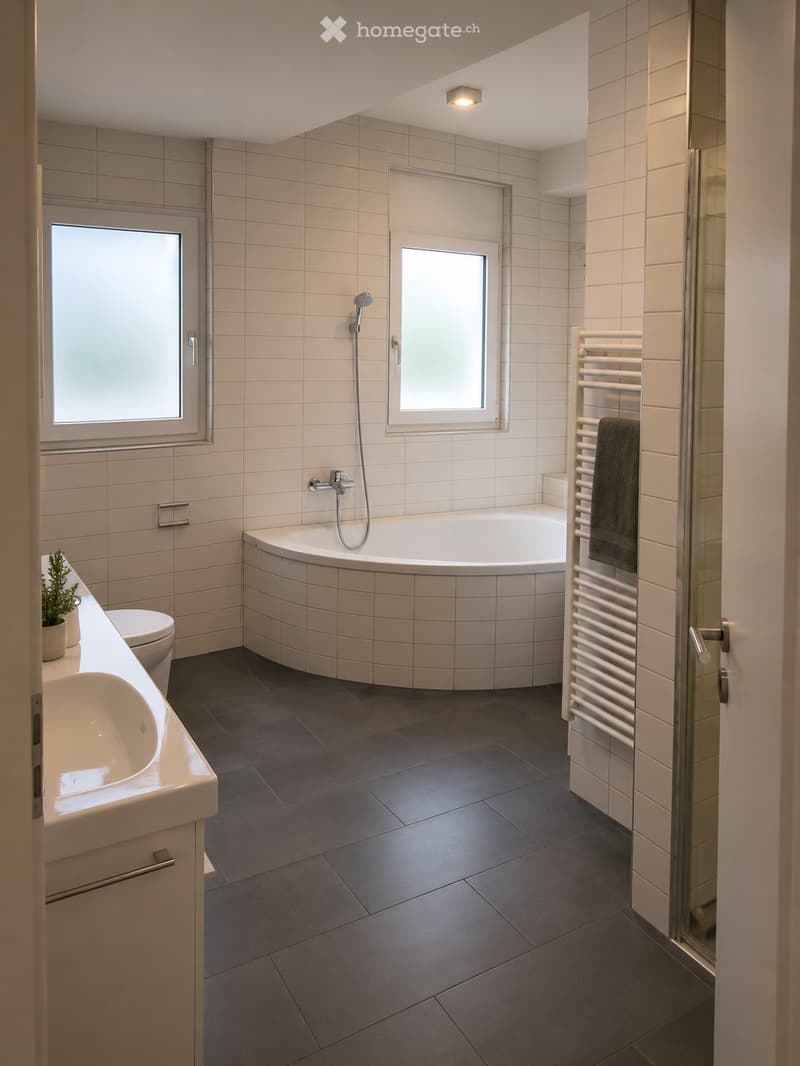Modern bathroom with a large bathtub, sink, toilet, radiator, and shower. Features tiled walls and dark tiled flooring.