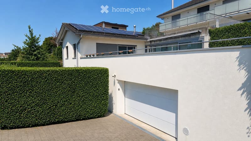 Modern two-story house with white exterior, solar panels on the roof, garage, and well-maintained hedges.