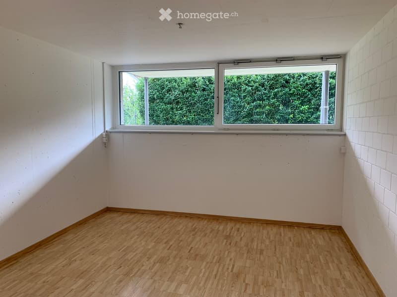 Empty room with wooden flooring, white walls, and a large window offering a view of greenery outside.