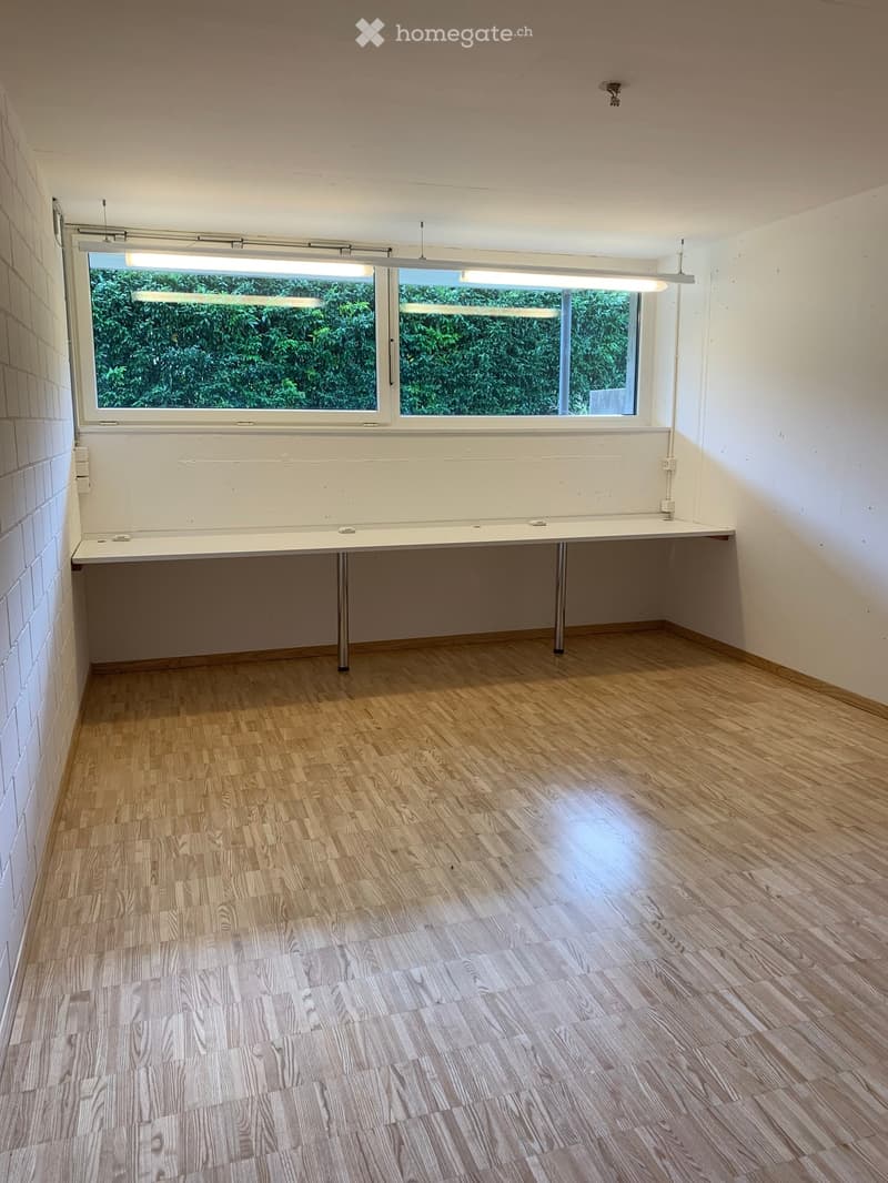 Empty room with wooden flooring, white walls, and a large window with a view of greenery. A simple table is positioned against one wall.