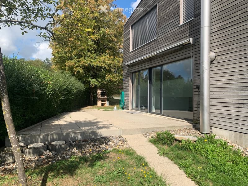 Modern wooden house with large glass sliding doors, raised patio, garden, and barbecue area.
