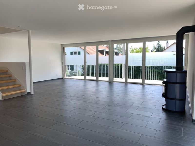 Empty room with dark grey tiled floor, white walls, large sliding glass doors leading to a balcony, wooden staircase, and a wood stove.