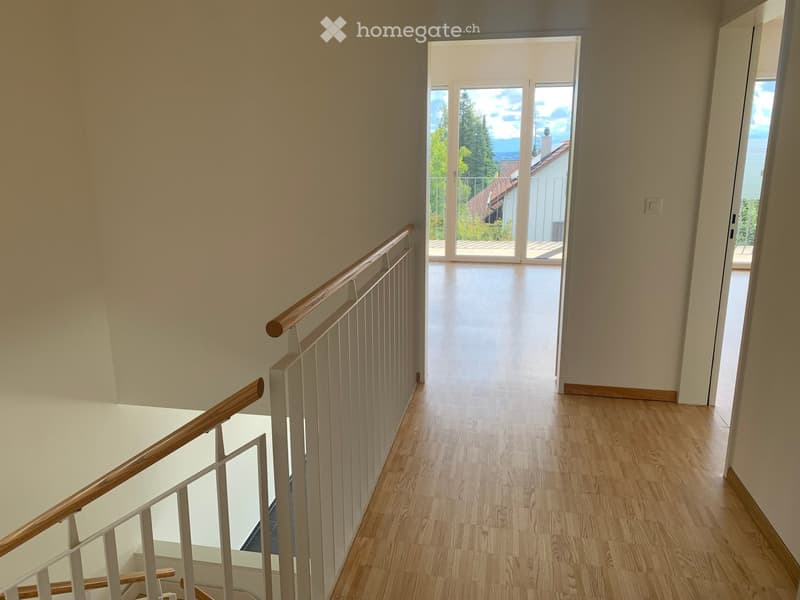 Spacious hallway with wooden flooring, white walls, and a wooden railing leading to an open area with large glass doors and a view of greenery.