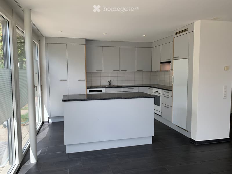 Modern kitchen with white cabinets, dark countertop, and built-in appliances including a sink, oven, and refrigerator. Large windows provide natural light.