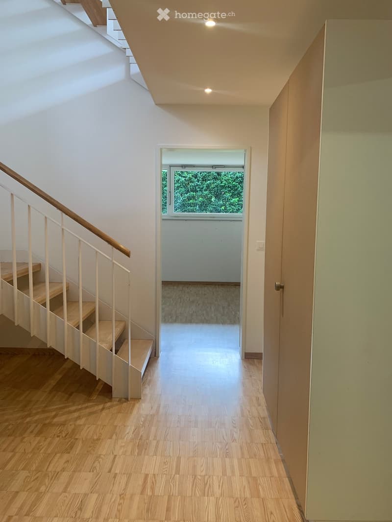 Bright hallway with wooden flooring, modern staircase with white railings, and a view to another room through an open doorway.