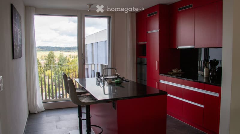 Modern kitchen with red cabinets, island, stools, and large windows offering a scenic view.