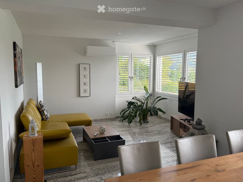 Modern living room with a yellow sectional sofa, coffee table, dining chairs, and a large plant near the window.
