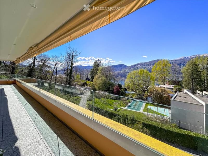 Balcony with glass railing, overlooking a scenic landscape with trees, a swimming pool, and distant mountains.