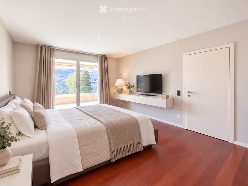 Modern bedroom with a large bed, pillows, blanket, table lamp, television, and potted plants. Large windows offer a scenic view.