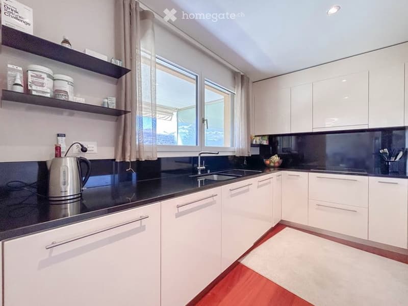 Modern kitchen with white cabinets, black countertops, and a stainless steel electric kettle on the counter. There is a window with a curtain, a sink with a faucet, and shelves with various items.