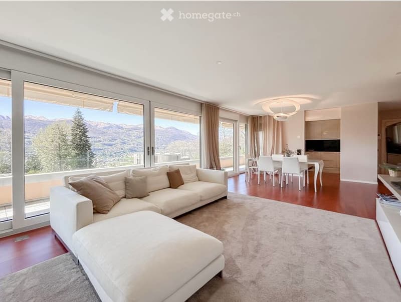 Modern living and dining area with large windows offering a scenic mountain view. Features a white sectional sofa with brown throw pillows, an ottoman, a dining table with chairs, and a sleek kitchen island.