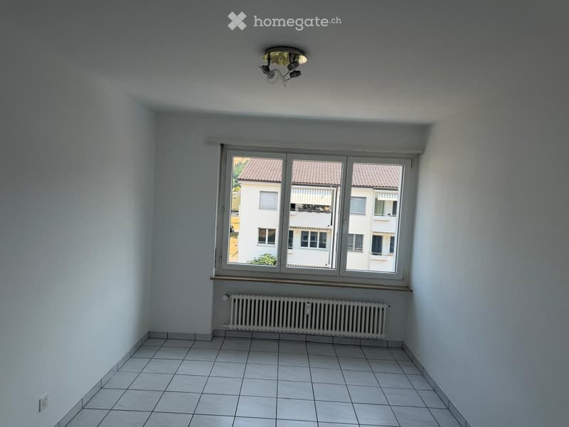 Empty room with tiled floor, white walls, large window with view of neighboring buildings, radiator, and ceiling light fixture.