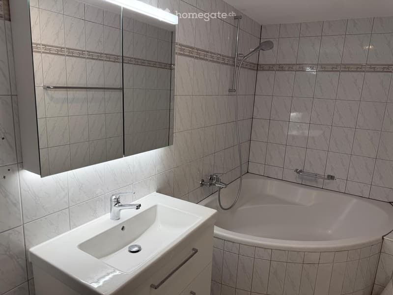 Modern bathroom with a white sink, chrome faucet, bathtub, and a large mirror with built-in lighting. The walls are tiled in a light marble pattern.