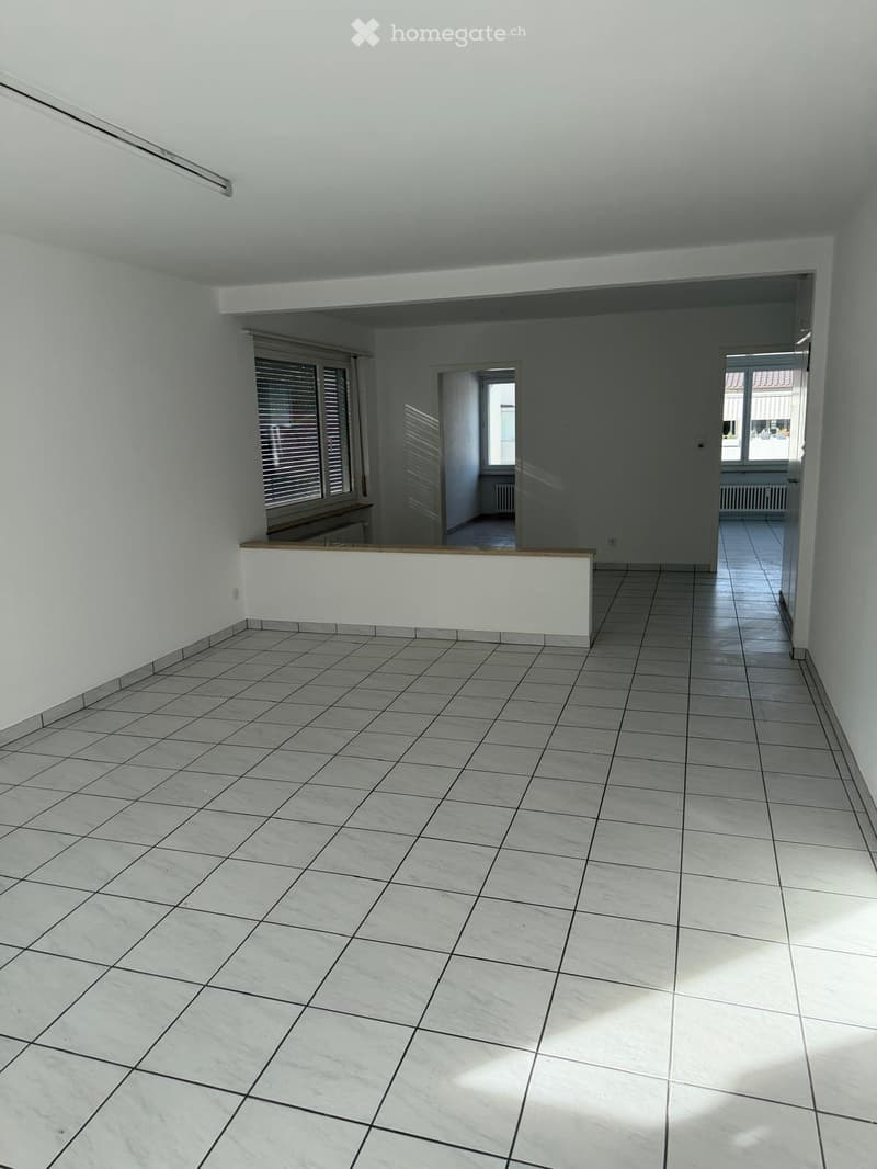 Empty room with tiled floor, white walls, and large windows with blinds. There is a white counter or partition in the center of the room.