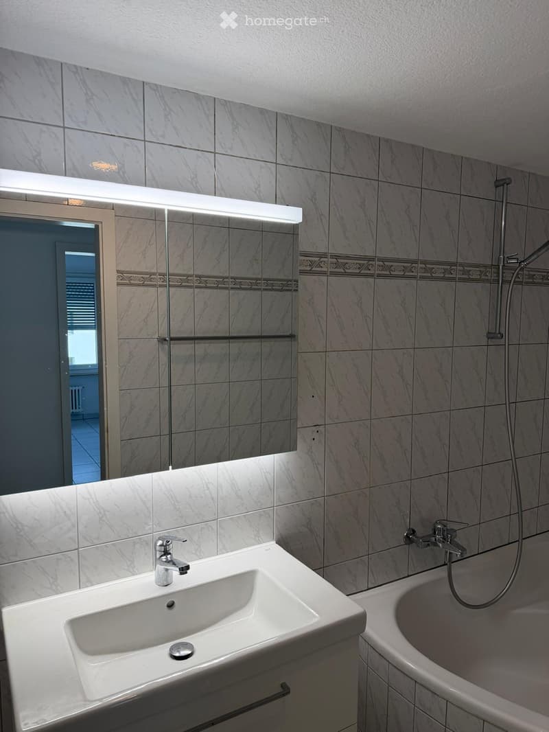 Modern bathroom with marble tiles, a white sink with a chrome faucet, a bathtub with a handheld showerhead, and a large mirror with built-in lighting.