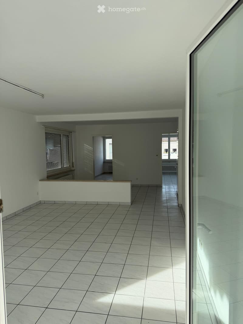 Empty hallway with tiled floor, white walls, and large windows with blinds. Natural light streams in from the windows, and the space appears clean and uncluttered.