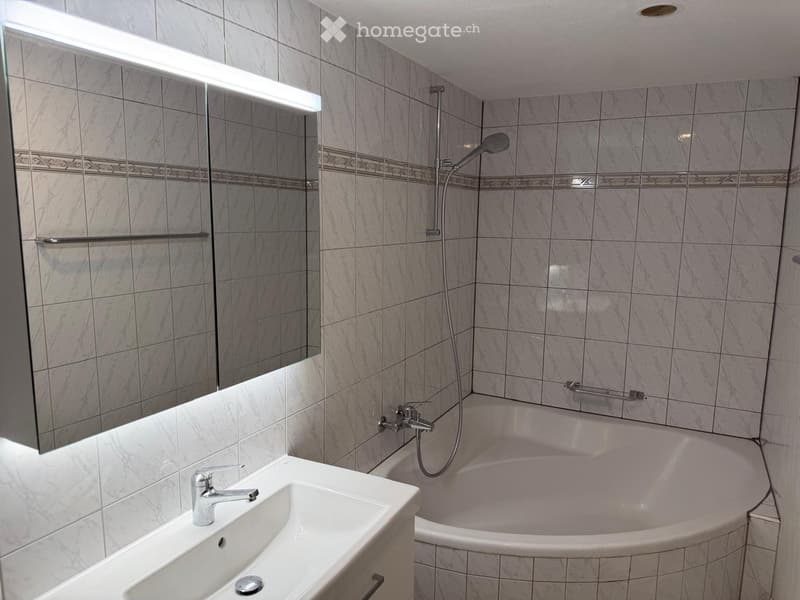 Modern bathroom with marble tiles, a white sink, a bathtub, a showerhead, and a large mirror with built-in lighting.