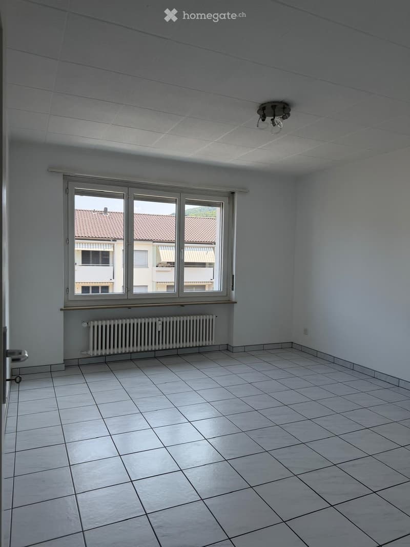 Empty room with white tiled floor, white walls, large window with view of neighboring building, radiator, and ceiling light fixture.