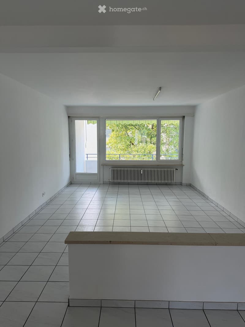 Empty room with white tiled floor, white walls, large windows, and a radiator. A wooden bench is placed in the foreground.