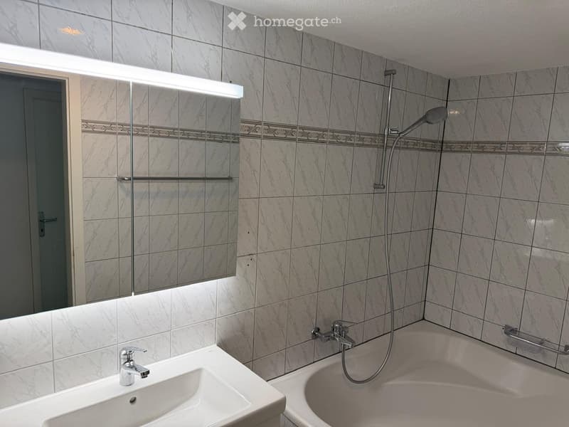Modern bathroom with marble tiles, a white sink with a chrome faucet, a bathtub with a handheld showerhead, and a large mirror with built-in lighting.