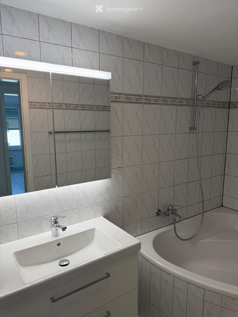 Modern bathroom with marble tiles, a white sink with a chrome faucet, a bathtub with a handheld showerhead, and a large mirror with built-in lighting.