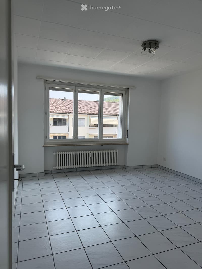 Empty room with white tiled floor, white walls, large window with view of neighboring building, radiator, and ceiling light fixture.