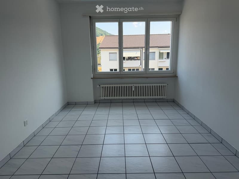 Empty room with white tiled floor, white walls, large window with view of neighboring buildings, radiator below window, and electrical outlet on wall.