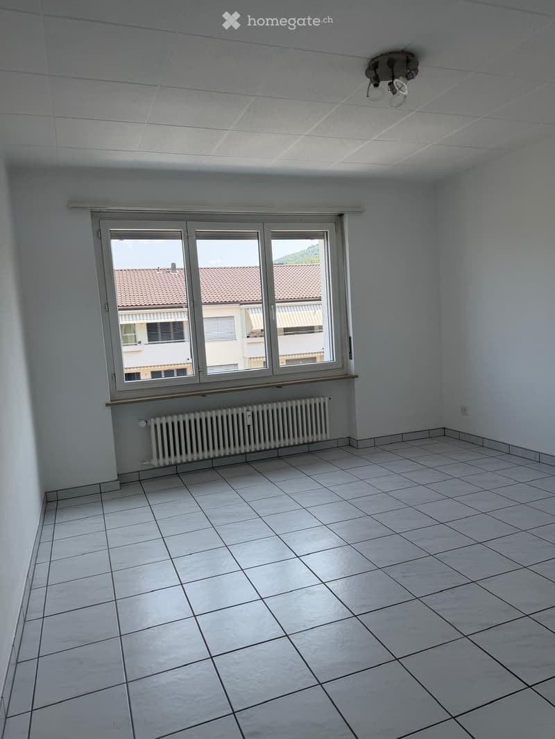 Empty room with white tiled floor, white walls, large window with view of neighboring building, radiator, and ceiling light fixture.