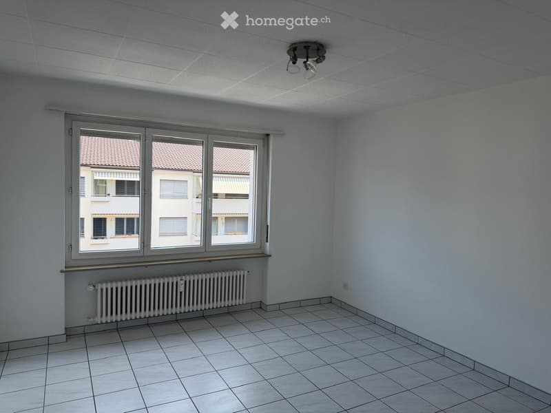 Empty room with tiled floor, white walls, large window with view of neighboring building, radiator, and ceiling light fixture.