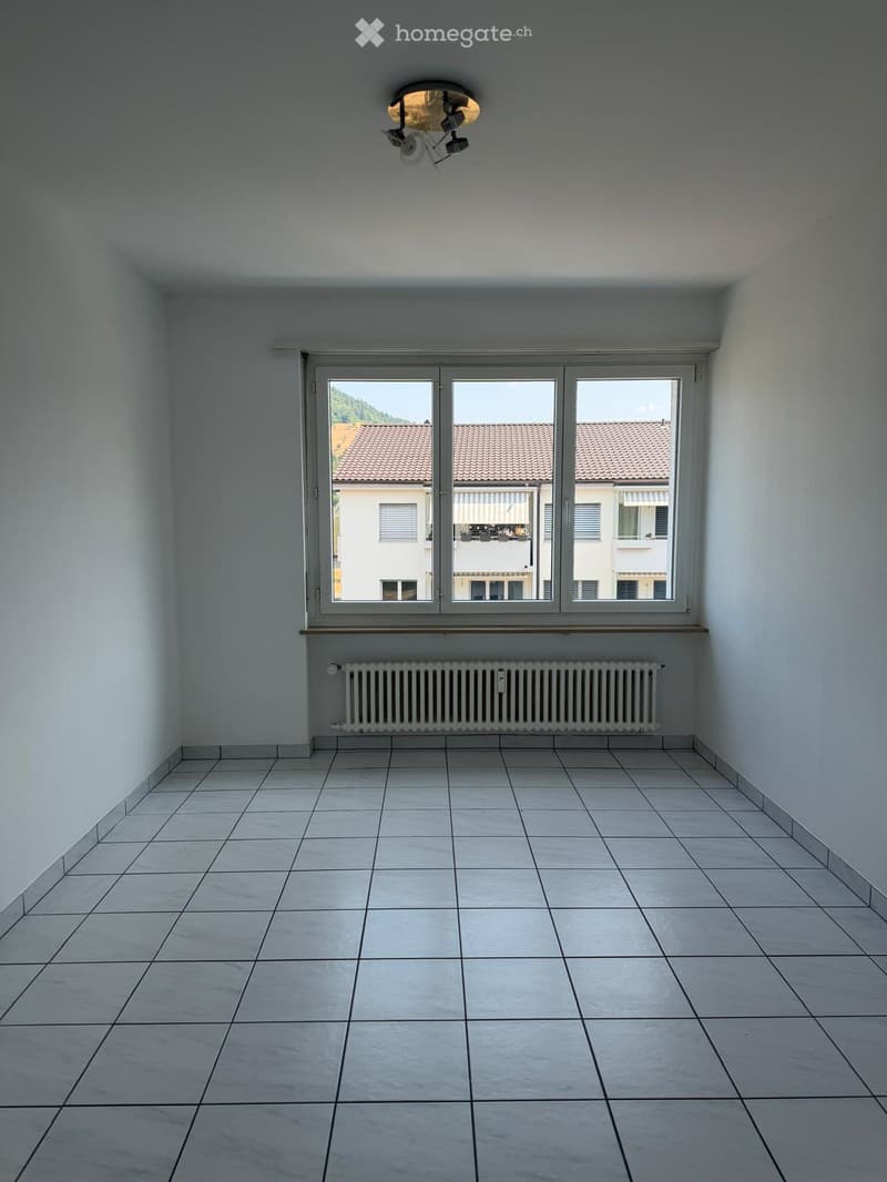 Empty room with tiled floor, white walls, large window with view of neighboring buildings, radiator, and ceiling light fixture.