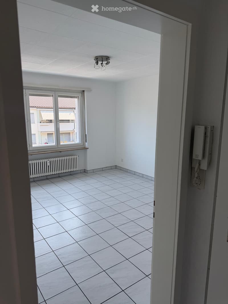 Empty room with white tiled floor, white walls, and a window with a radiator below it. The room has a ceiling light fixture and a telephone mounted on the wall.