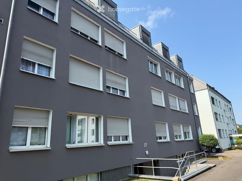Four-story residential building with grey facade, multiple windows with white shutters, and a small staircase leading to the entrance.