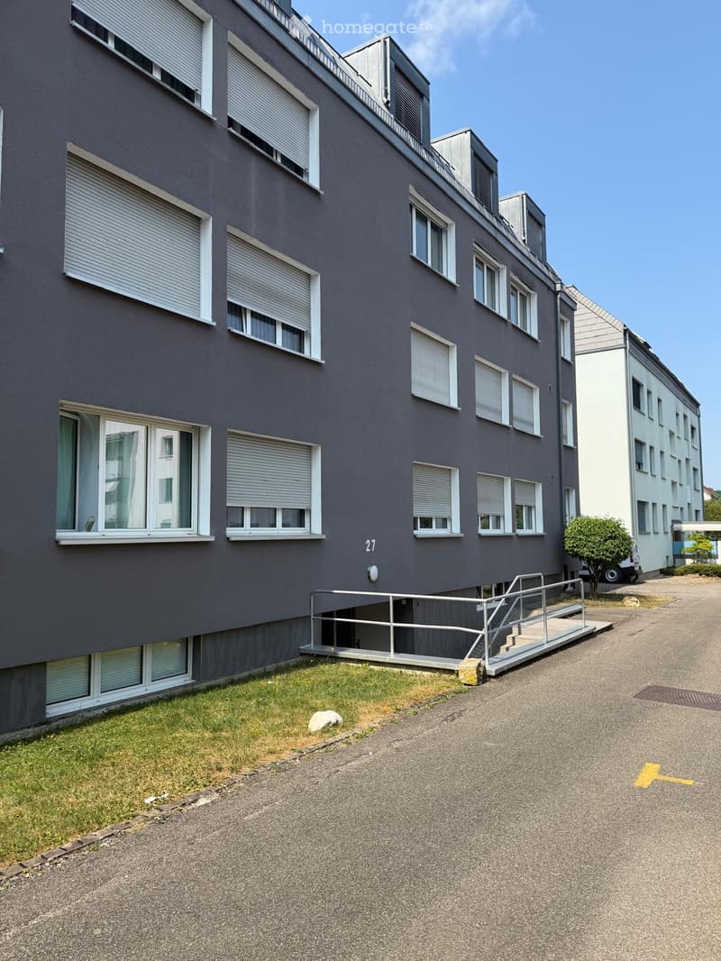A multi-story residential building with grey exterior walls, multiple windows with white frames, and a small staircase leading to the entrance.