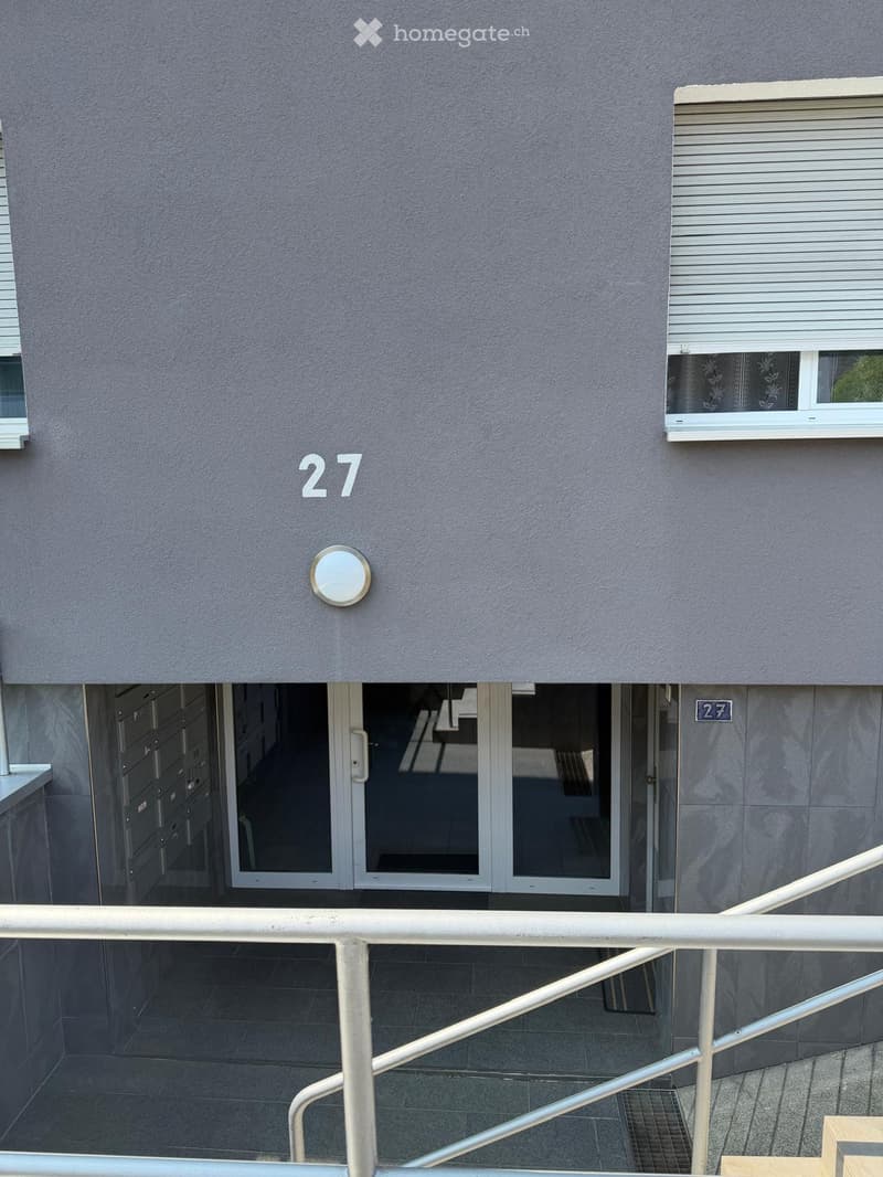 Exterior view of a multi-story building with grey facade, number 27 displayed prominently, a small round light fixture, and a glass entrance door with a metal railing leading up to it.