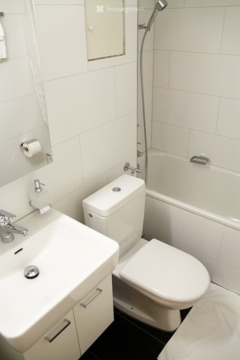 Modern bathroom with white tiles, a sink, toilet, bathtub, and showerhead. Includes a mirror and toilet paper holder.