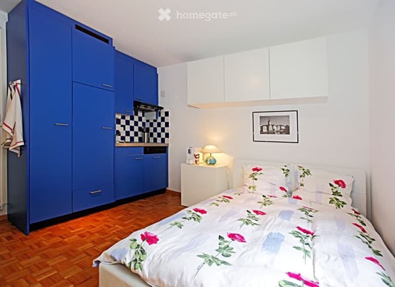 A compact studio apartment with a bed, nightstand, lamp, blue cabinets, and a picture frame on the wall.