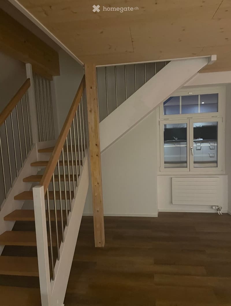 Empty hallway with wooden stairs, metal railing, and a window with glass panels. The floor is dark, and the walls are light-colored.