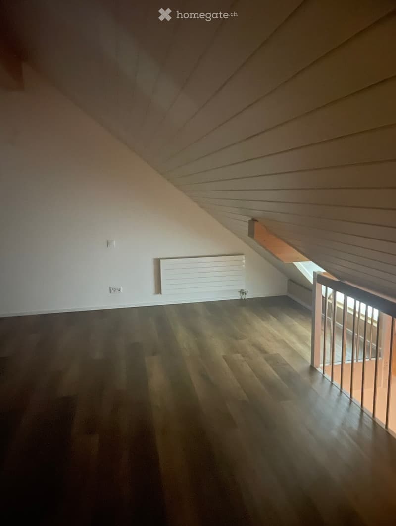 An empty attic space with wooden flooring, white walls, and a slanted wooden ceiling. A small window allows natural light to enter, and a white radiator is mounted on the wall.