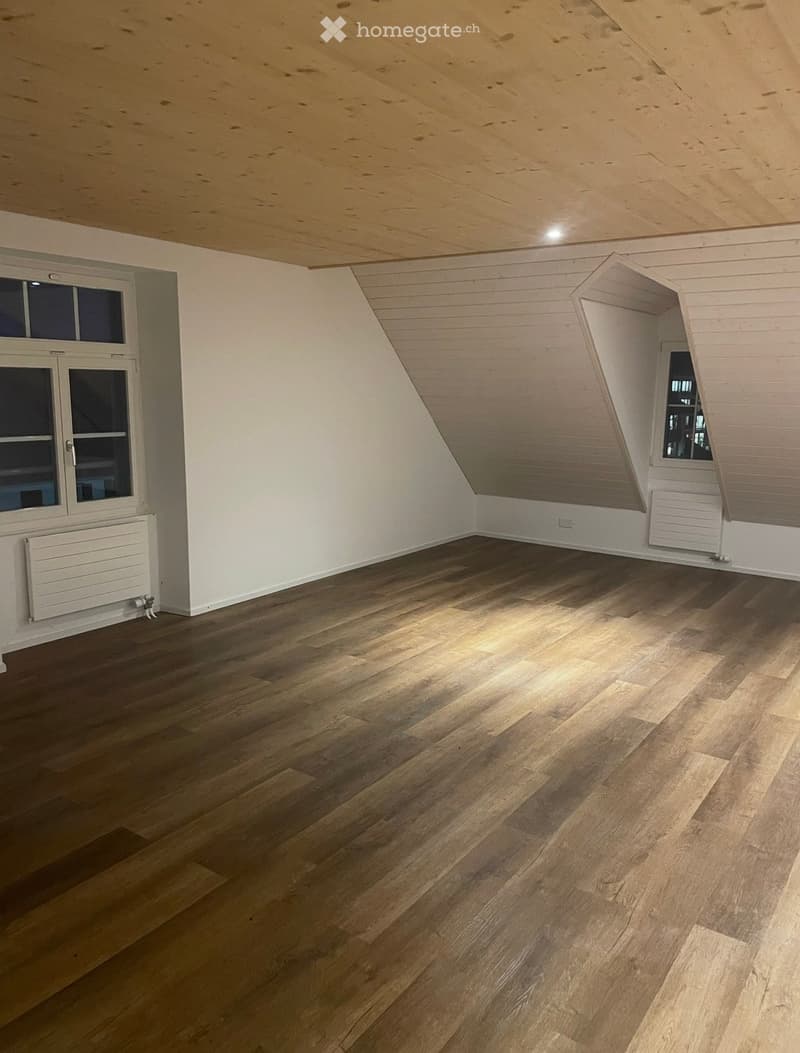 Empty attic room with wooden flooring, white walls, exposed wooden ceiling beams, and windows on two walls.