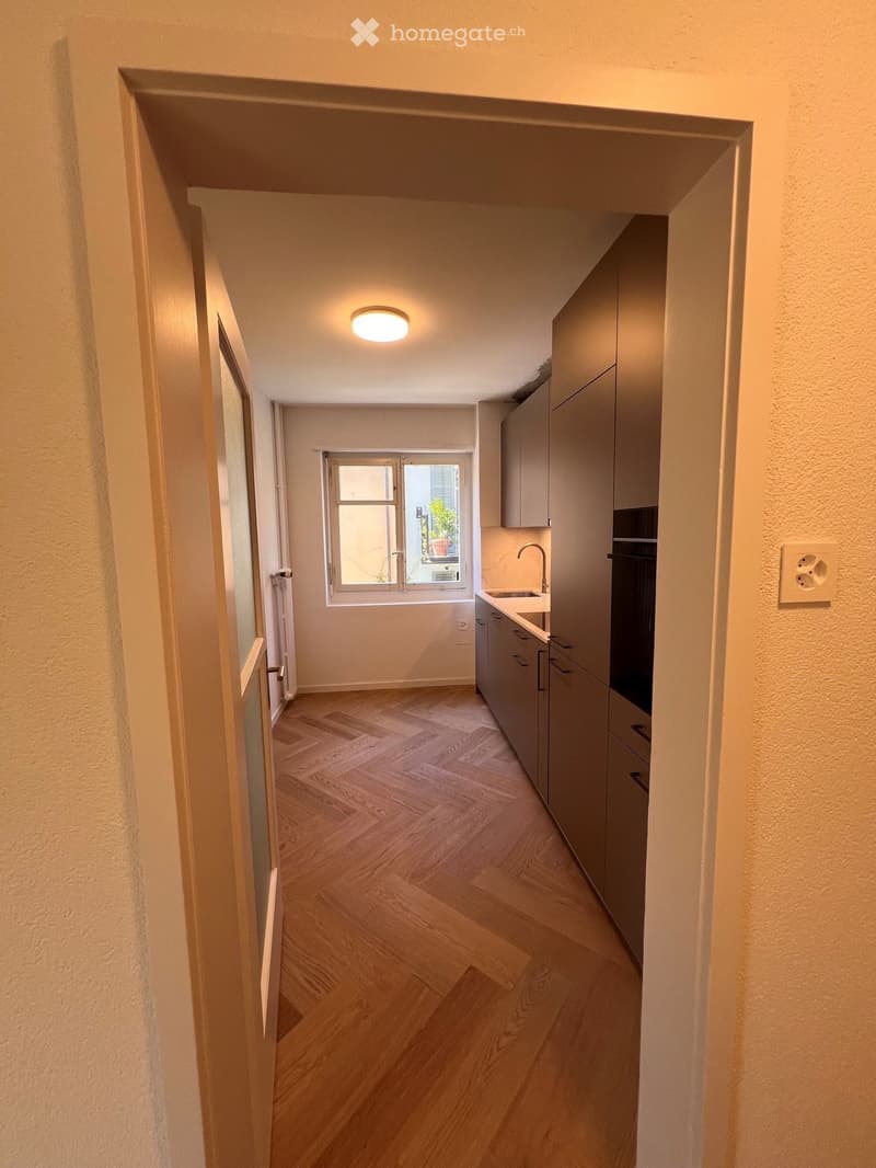 A narrow hallway with wooden flooring, a ceiling light, and a small window at the end. The hallway leads to a kitchen area with cabinets, a sink, and a stove.