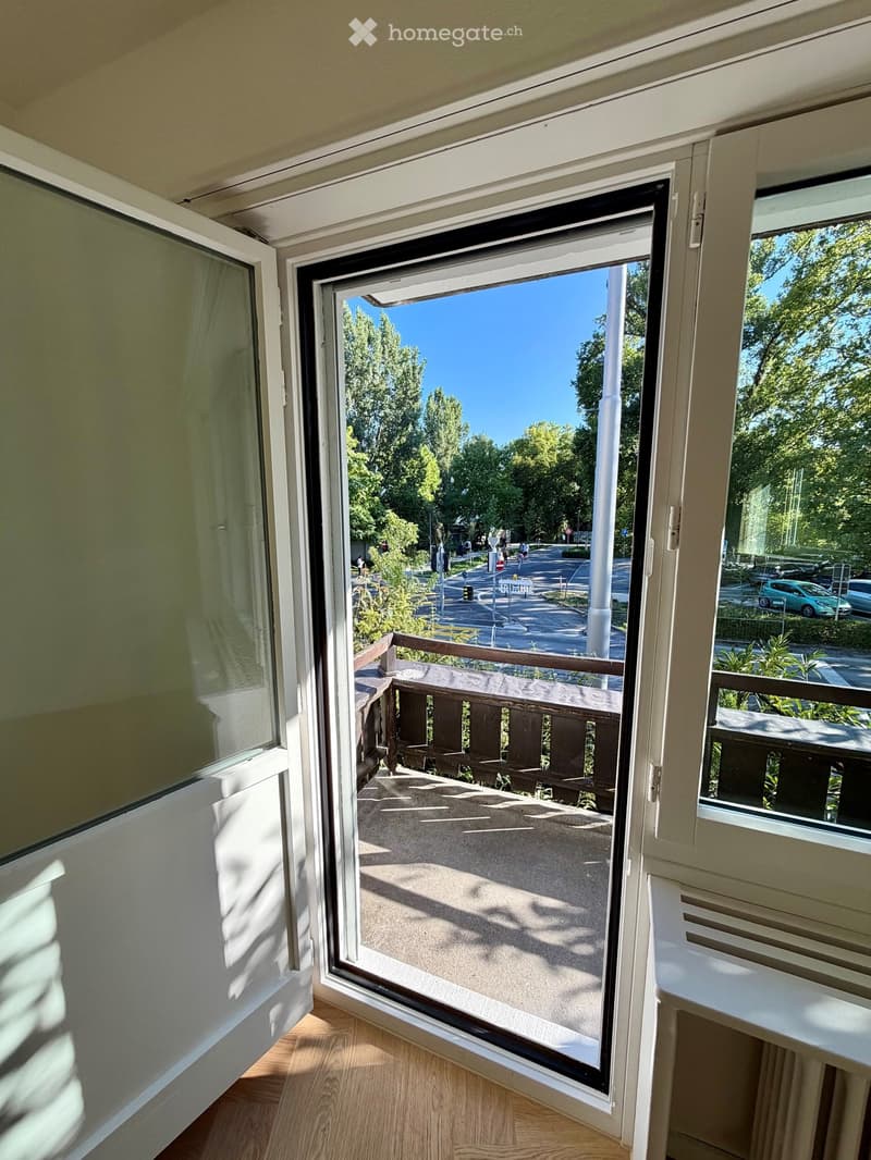 Open sliding window leading to a small balcony with wooden railing, overlooking a street with trees and parked cars.
