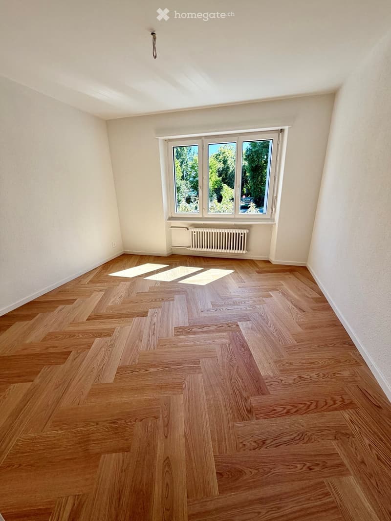 Empty room with wooden flooring, white walls, and a large window with a view of greenery. A radiator is visible below the window.