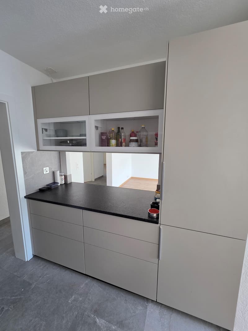 Modern kitchen with grey cabinets, black countertop, and open shelving displaying bottles and a pan. The floor is tiled, and the space is well-lit with natural light from the adjacent room.